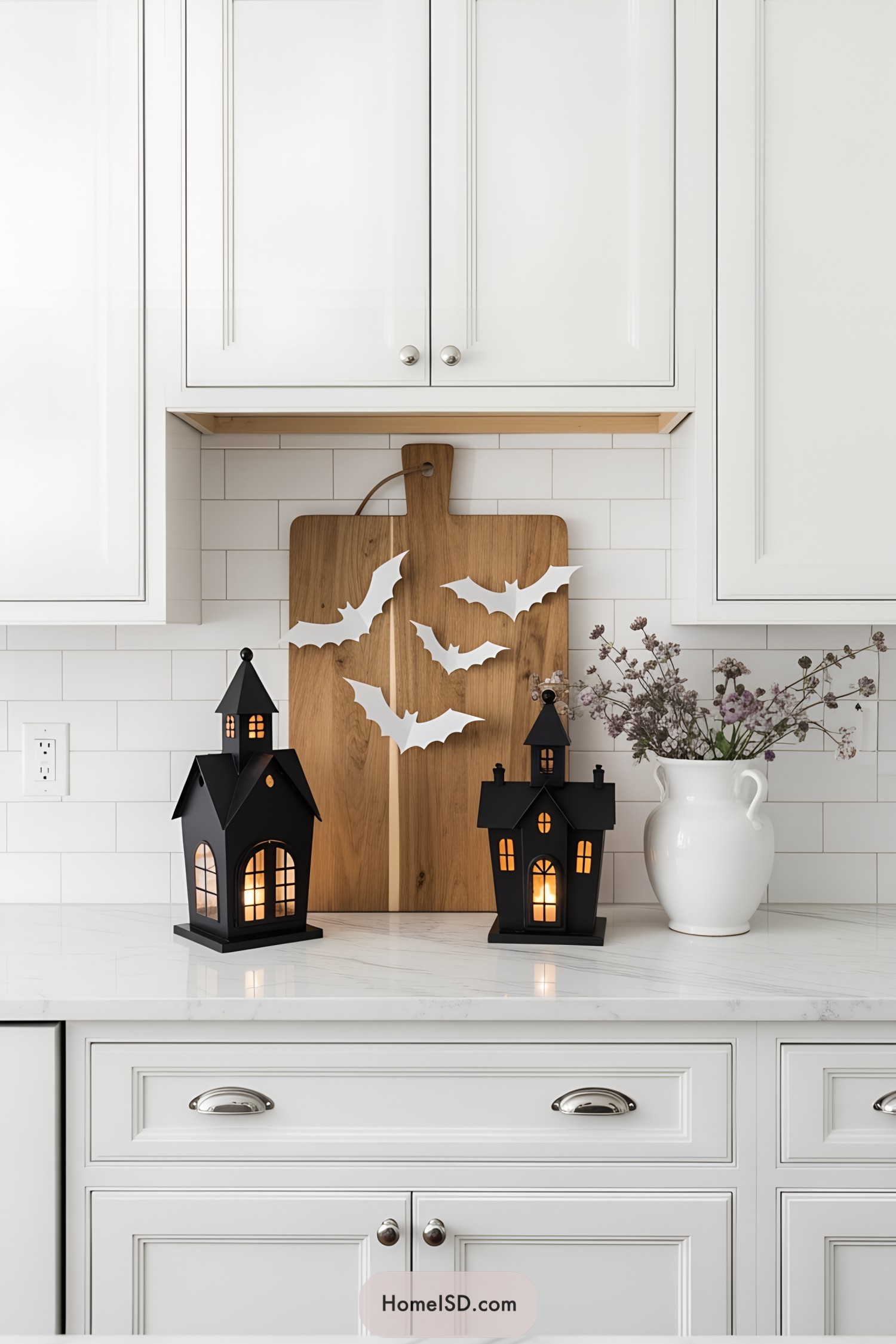 Elegant Halloween decor idea with a bright kitchen setting, featuring a clean white color theme and classic cabinetry. The walls display white subway tiles, and countertops are polished white marble. Upper and lower cabinets have raised panel doors with chrome knobs. Decor includes a wooden cutting board as backdrop, matte black metal lanterns shaped like haunted houses with warm interior lighting, and delicate paper bats for a subtle spooky accent. A white ceramic vase with muted purple floral arrangements adds sophisticated contrast. The overall aesthetic remains minimal, refined, and slightly rustic.