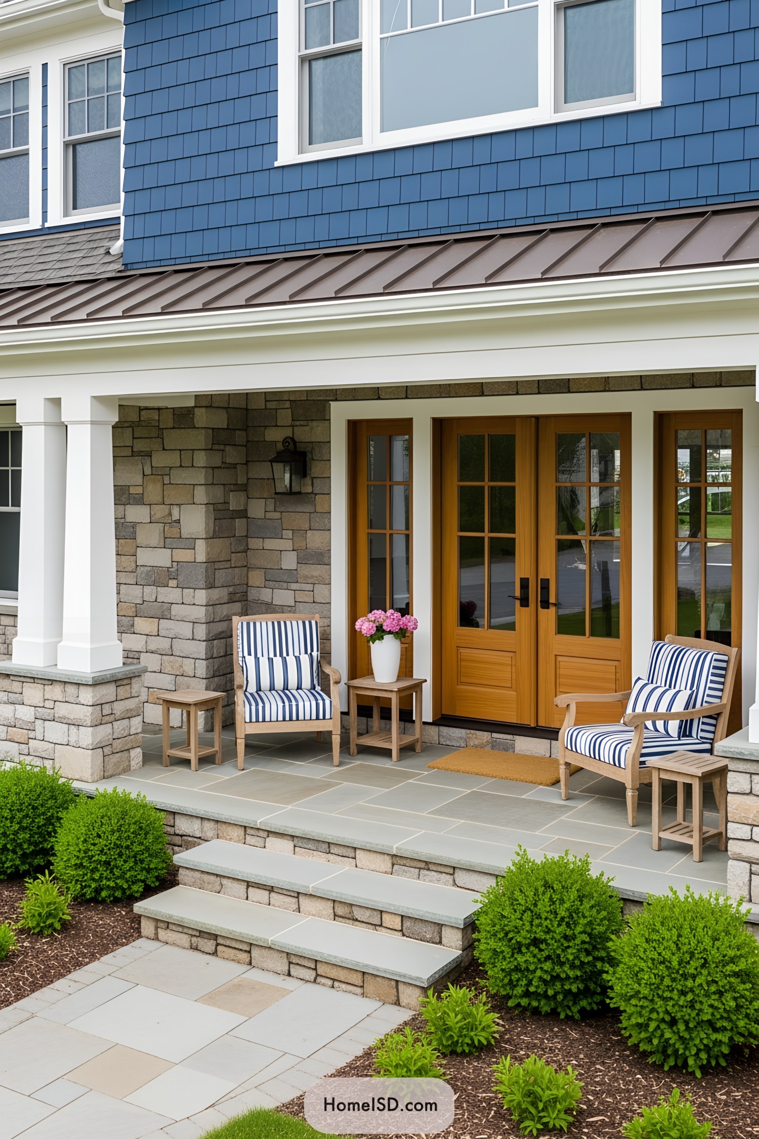 Front porch curb appeal with a classic-coastal aesthetic, featuring a navy blue shingled facade and natural stone accents. The structure displays warm wood-framed double doors and white trim. The porch floor and steps are constructed from flagstone, complemented by a partial stone wall. Two wooden armchairs with blue-and-white striped cushions flank matching side tables, one holding a white vase with pink flowers. Lush green shrubs and neatly mulched garden beds create a welcoming landscape in front of the porch.