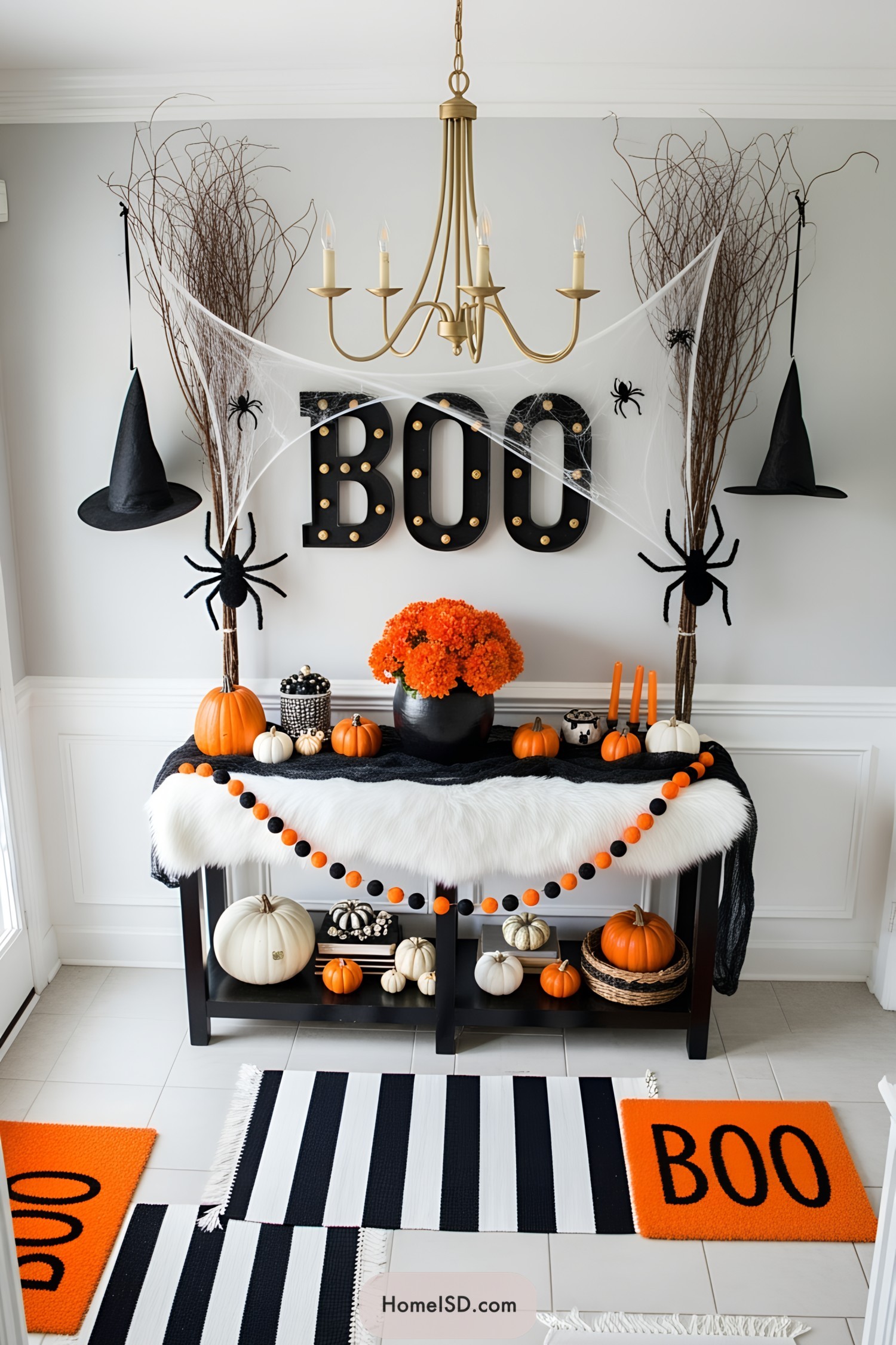 Halloween entryway with whimsical, eclectic style and a festive, monochrome color palette of black, orange, and white. Light gray walls and white trim create a crisp backdrop. The floor is neutral tile with black and white striped rugs and an orange “BOO” mat. A gold chandelier hangs from the plain white ceiling. The console features a faux fur white table cover, black gauze runner, assorted pumpkins, bright orange floral arrangement in a black pot, and a garland of orange and black pom-poms. Tall twigs and faux spiderwebs with large spiders frame a large “BOO” sign. Suspended black witch hats add a playful touch. No windows or view present.