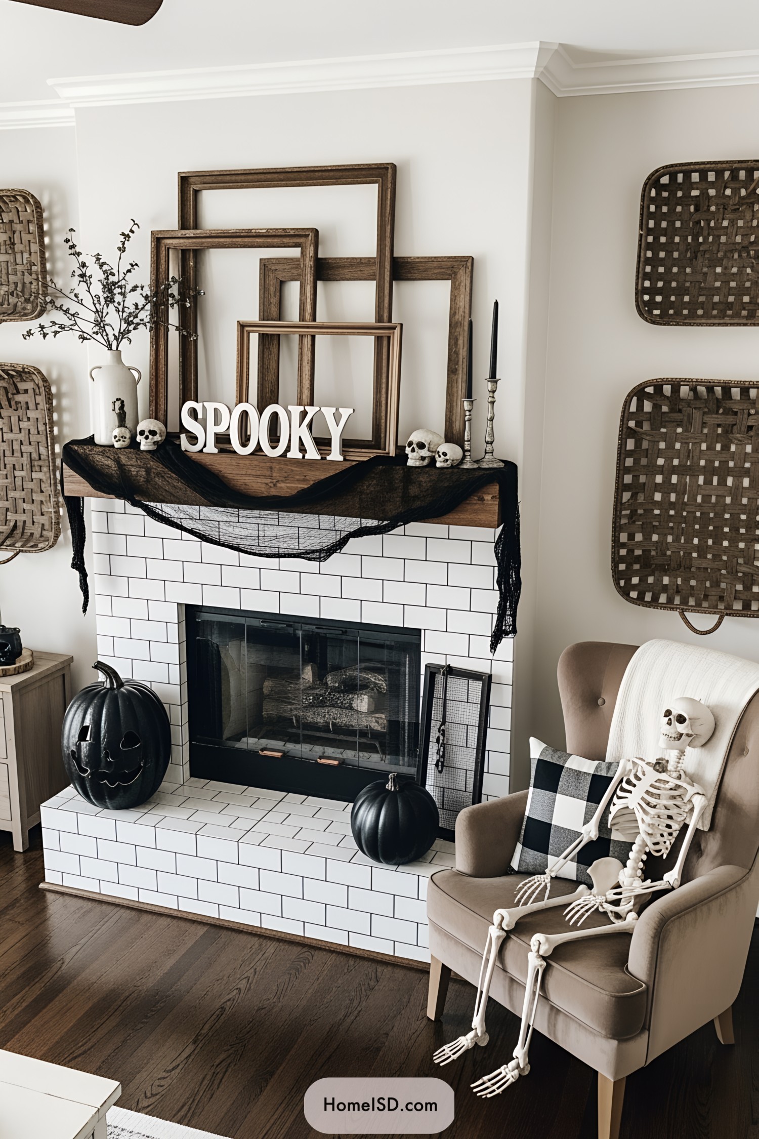 Living room with a cozy, rustic Halloween theme and farmhouse style. Color palette is neutral with black and white accents, featuring dark wood and creamy walls. The fireplace mantle is decorated with layered vintage picture frames, a “spooky” sign, black gauze, and small skull accents. White subway tile surrounds the fireplace. The walls are adorned with woven baskets. Dark wood flooring grounds the space. A plush, brown velvet armchair with a buffalo check pillow sits beside the hearth, holding a skeleton. A matte black pumpkin adds a whimsical touch. No windows are visible, and accessories utilize natural and vintage materials for a chic, eerie look.