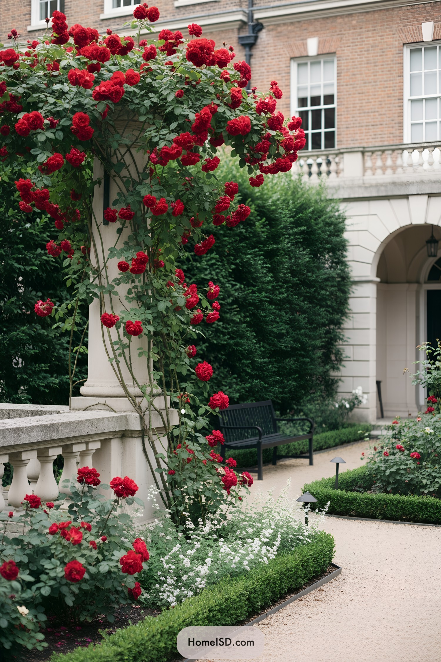 Red climbing roses drape a balustrade beside clipped hedges and a gravel path