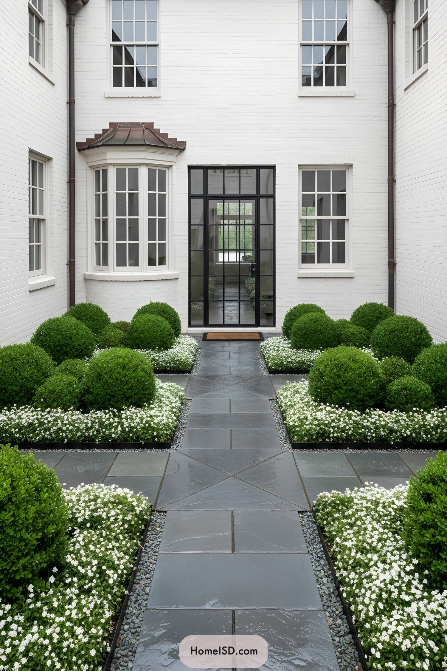 Symmetrical courtyard with round boxwood mounds, white flowers, and a slate walkway against white brick walls