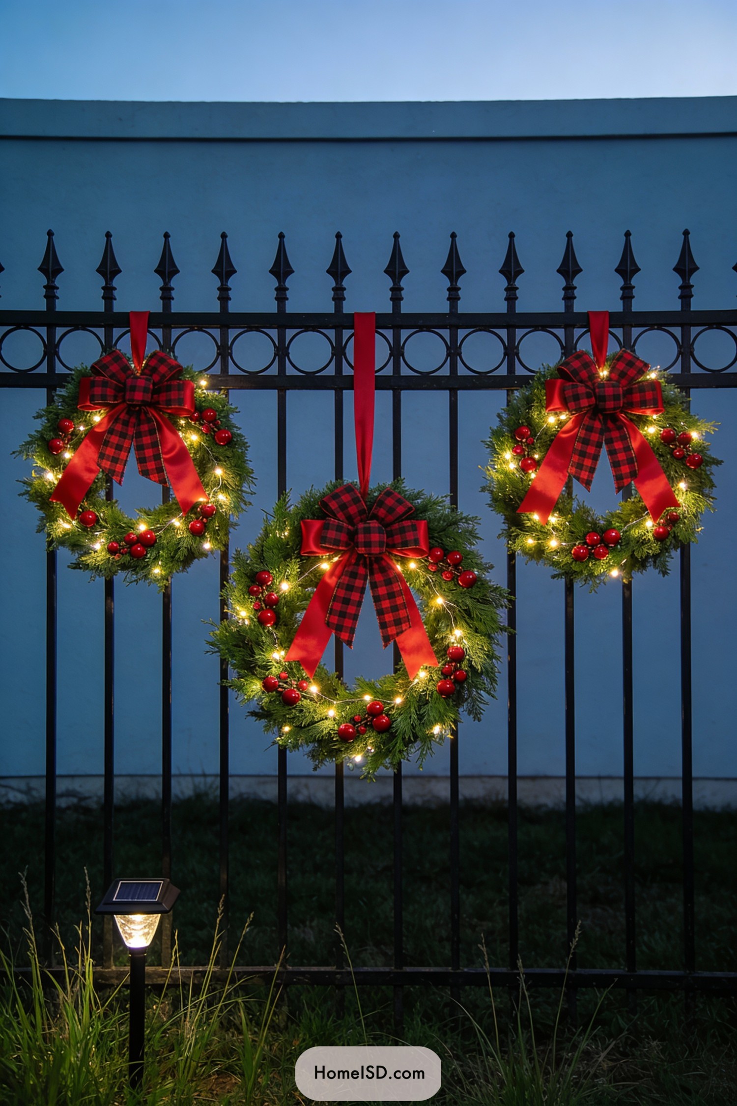 Three lighted Christmas wreaths with plaid bows hanging on a black iron fence at dusk