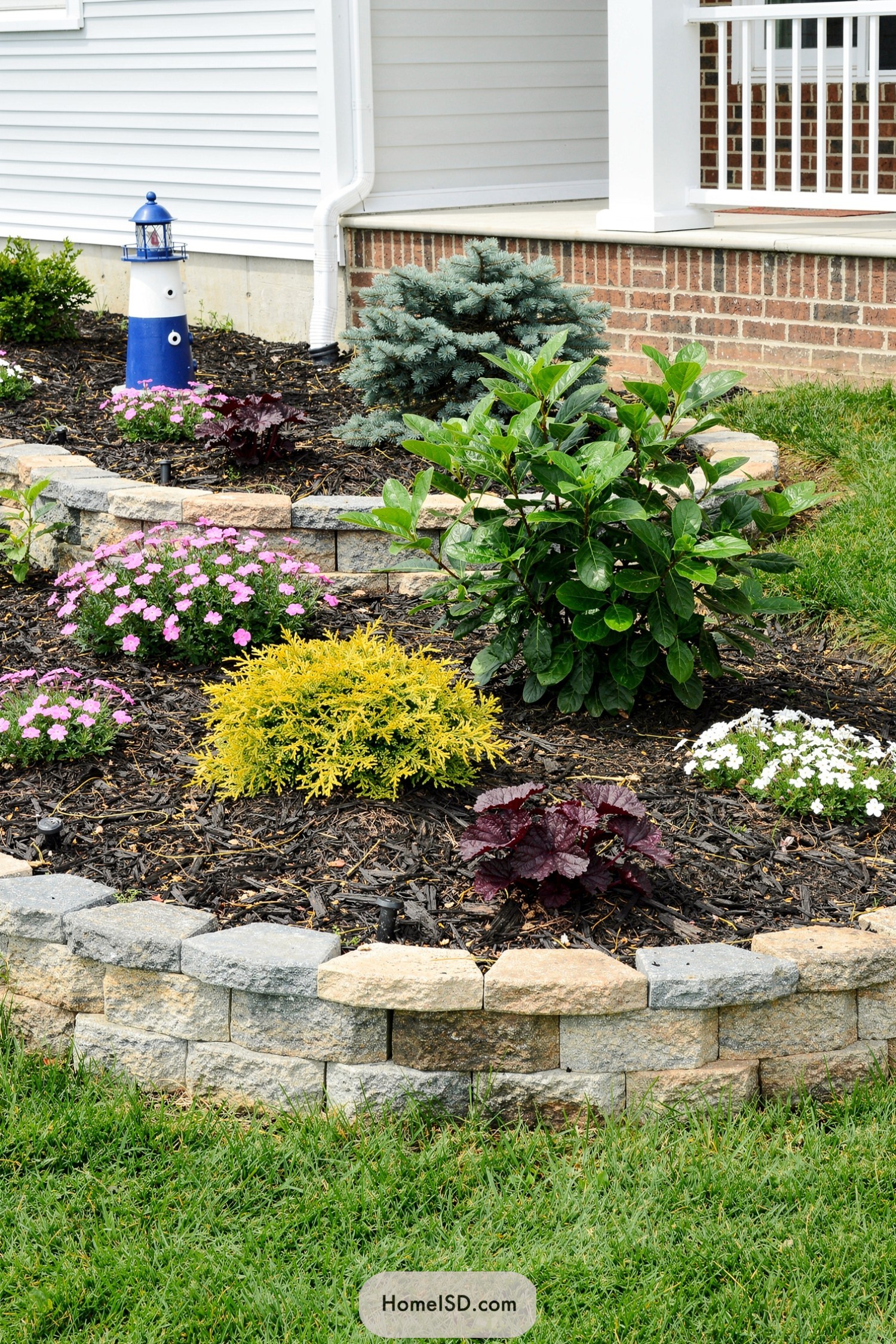 Curved stone-edged front flower bed with mixed shrubs, blooms, mulch, and a small blue-and-white lighthouse ornament