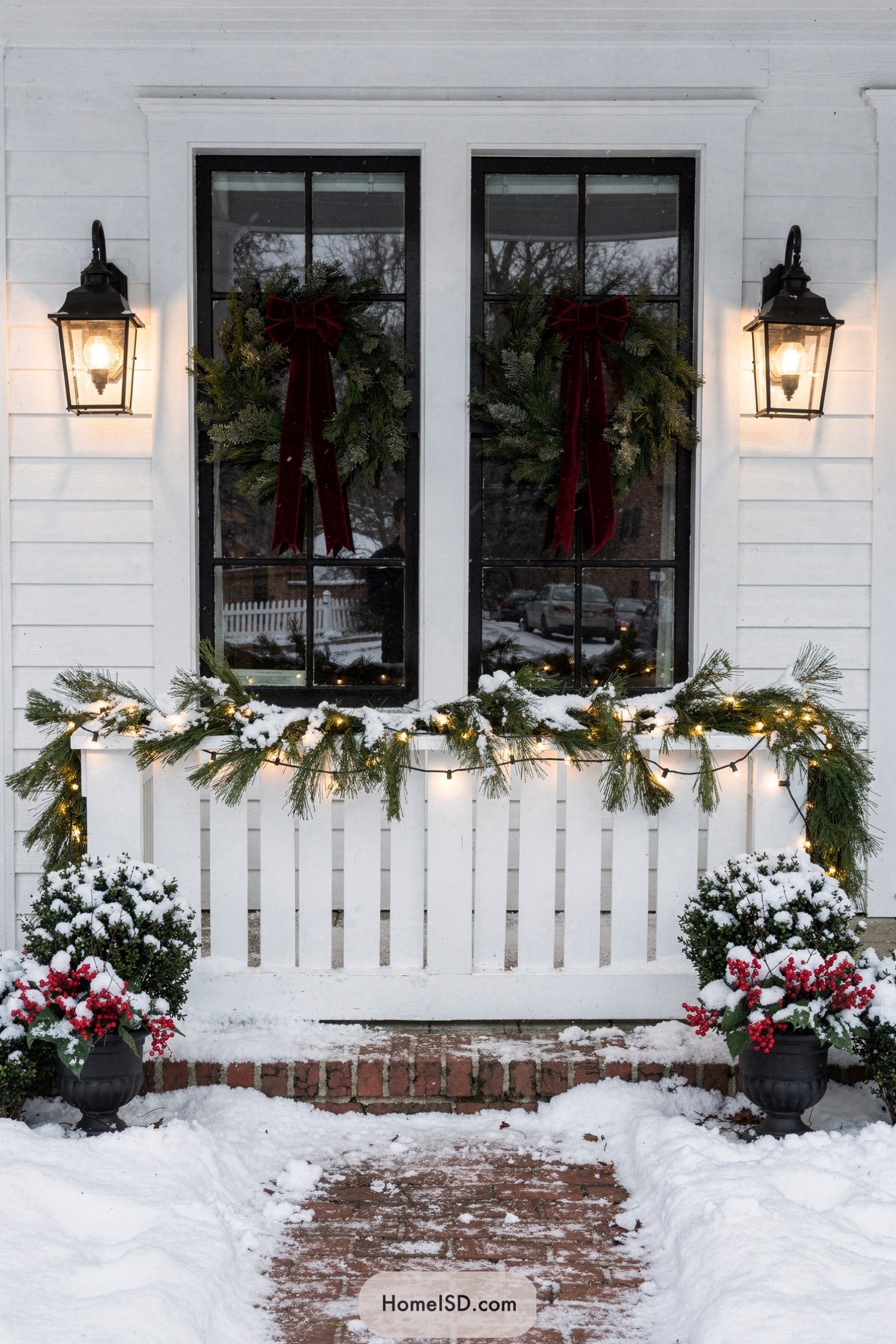 Snowy front porch with lit evergreen wreaths, garland, and lanterns framing twin cottage windows