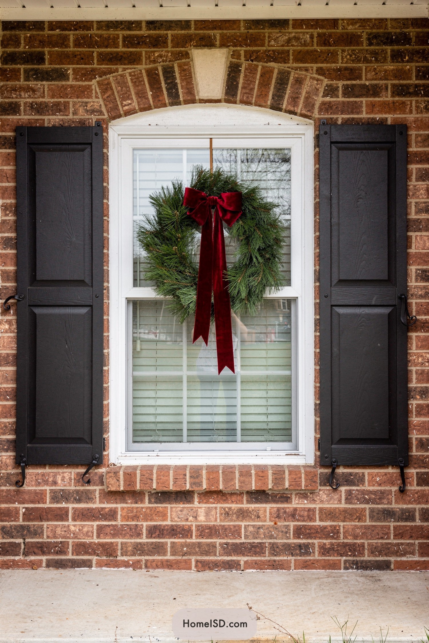 Green wreath with long red velvet bow hanging in a white-trimmed window on a brick house with black shutters