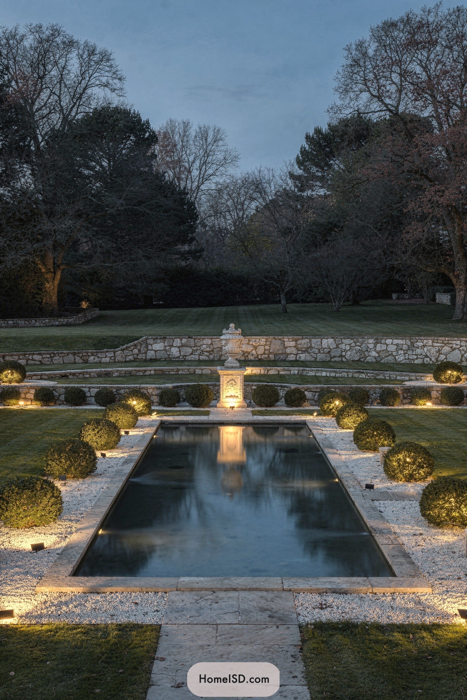 Formal garden pool flanked by lit boxwoods and central stone statue at dusk