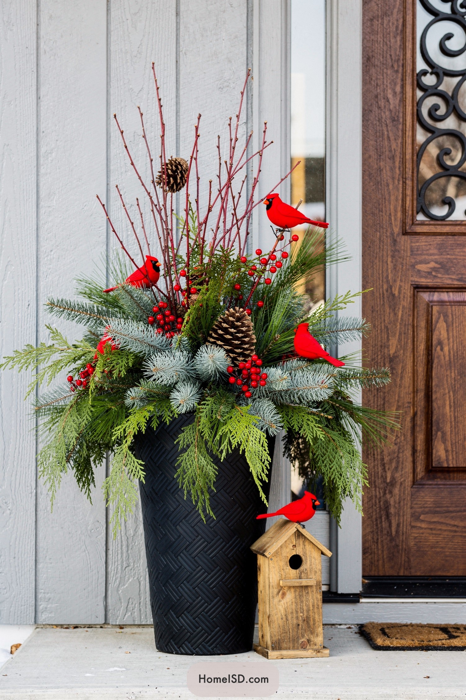Tall black planter filled with mixed evergreens, red berries, twigs, pinecones, and bright red cardinal accents beside a small wooden birdhouse at a front door