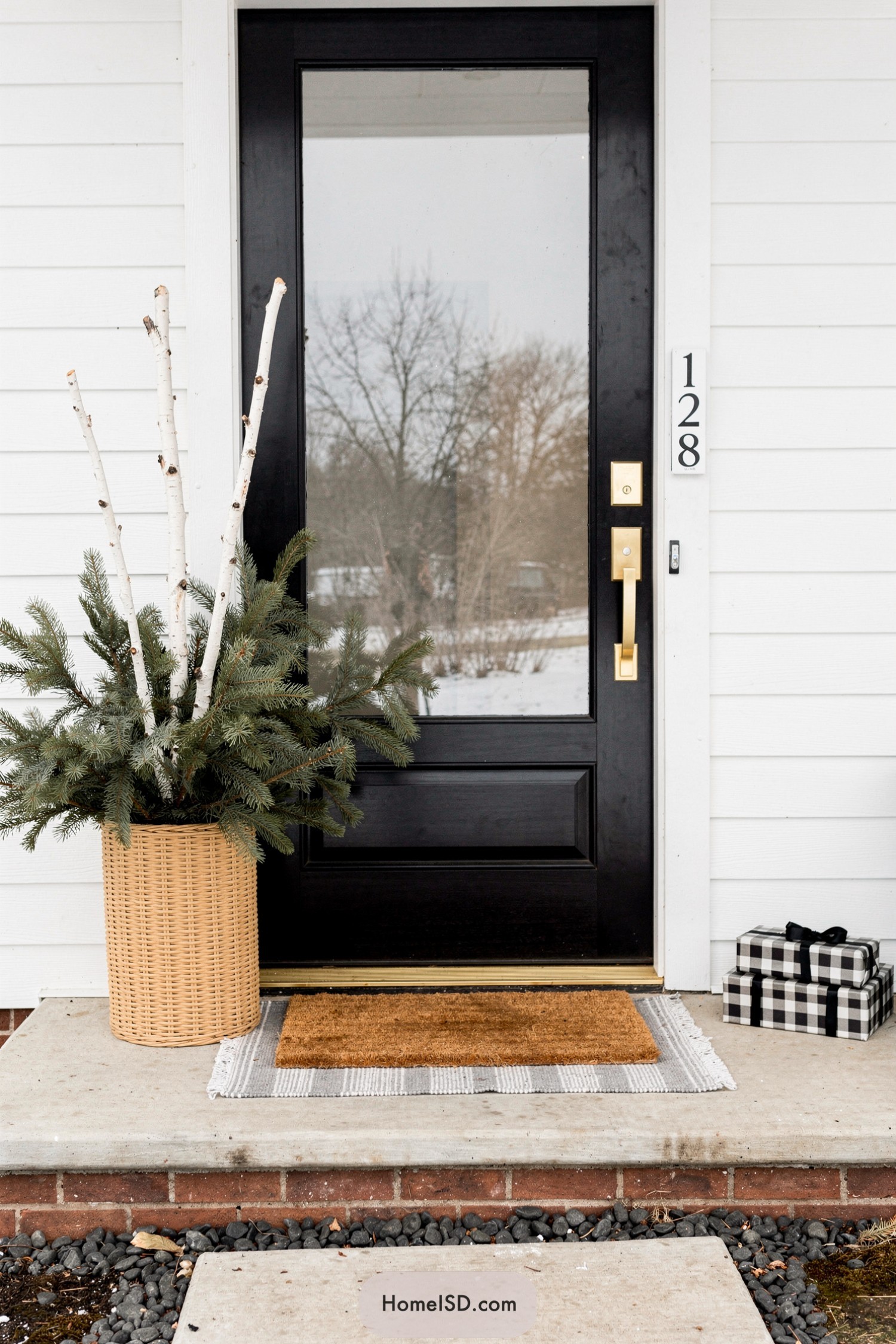 Tall birch branches and evergreen boughs in a woven basket planter beside a modern black front door with layered doormats and buffalo check gift boxes