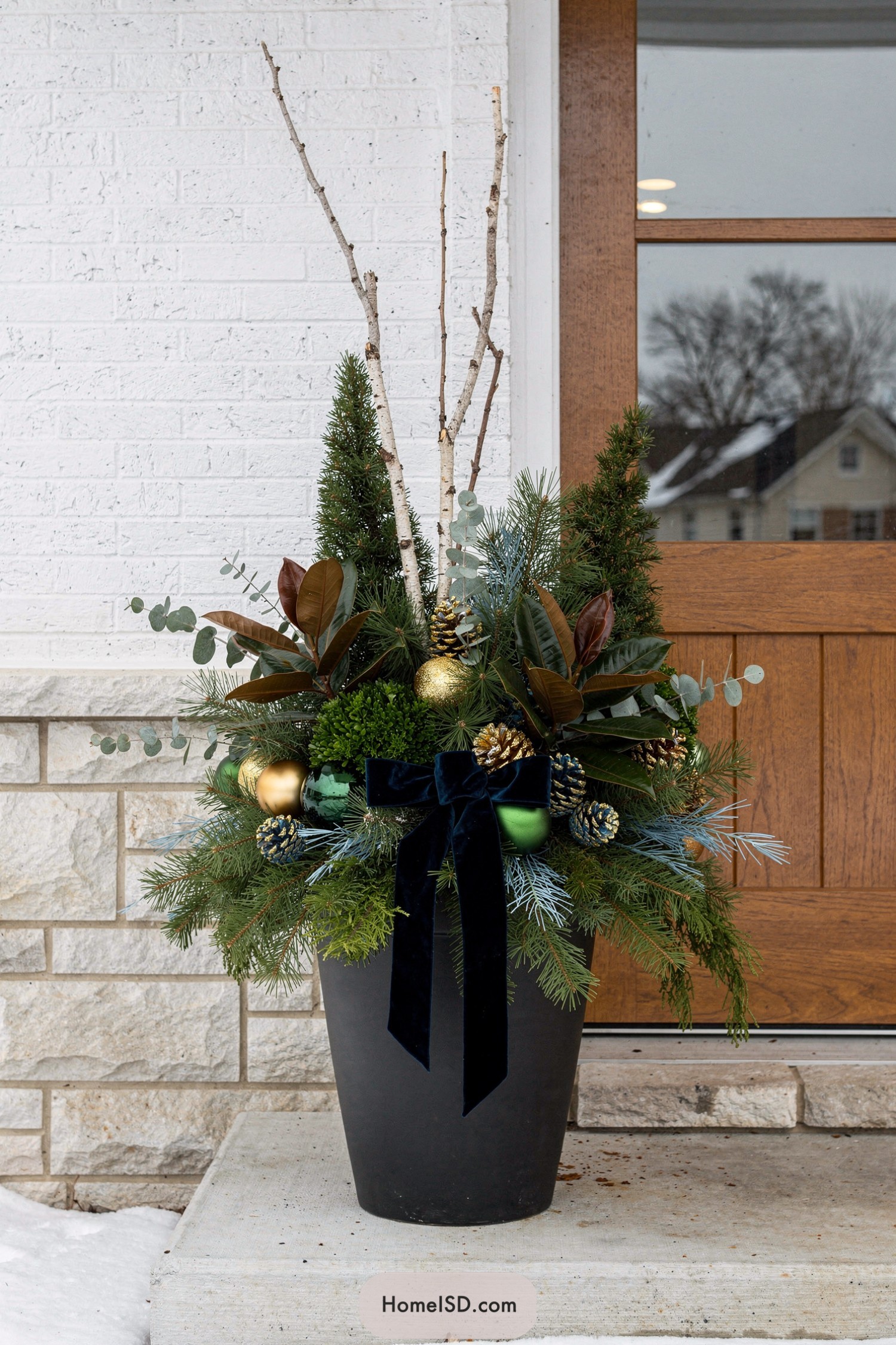 Tall black planter filled with winter evergreens, birch branches, ornaments, and a navy velvet bow by a front door