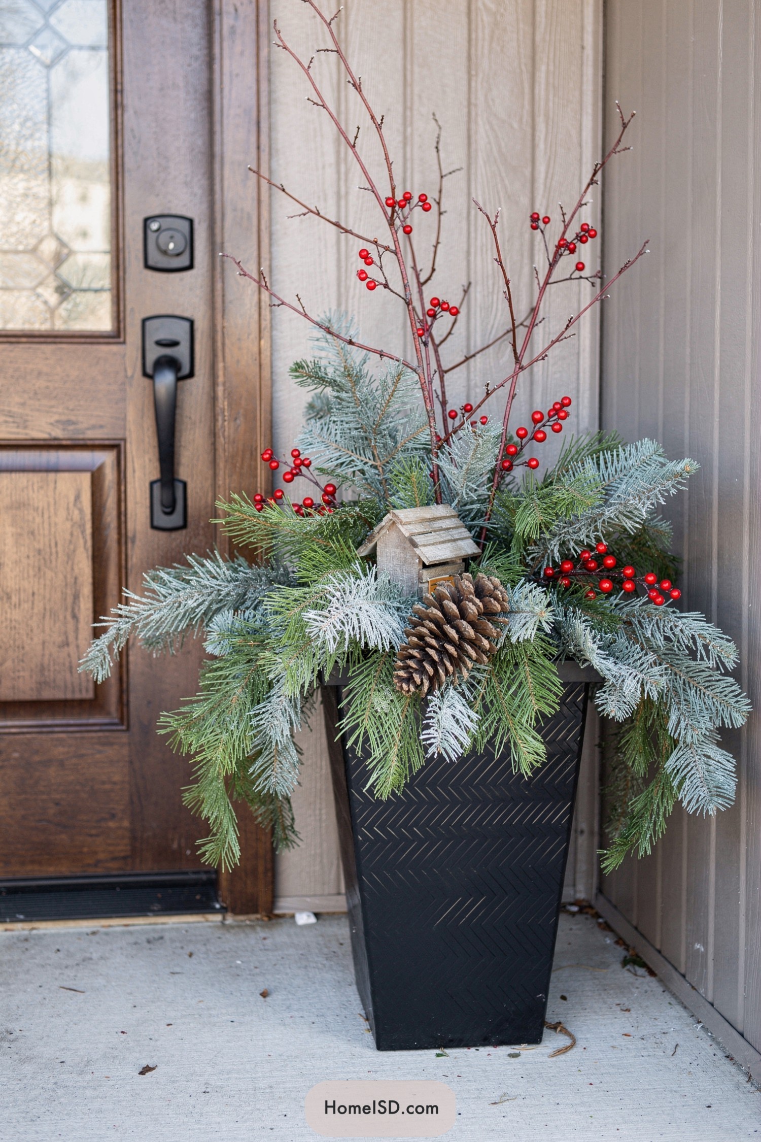 Tall black planter with frosted evergreens, red berries, birdhouse, and pinecone by a wood front door