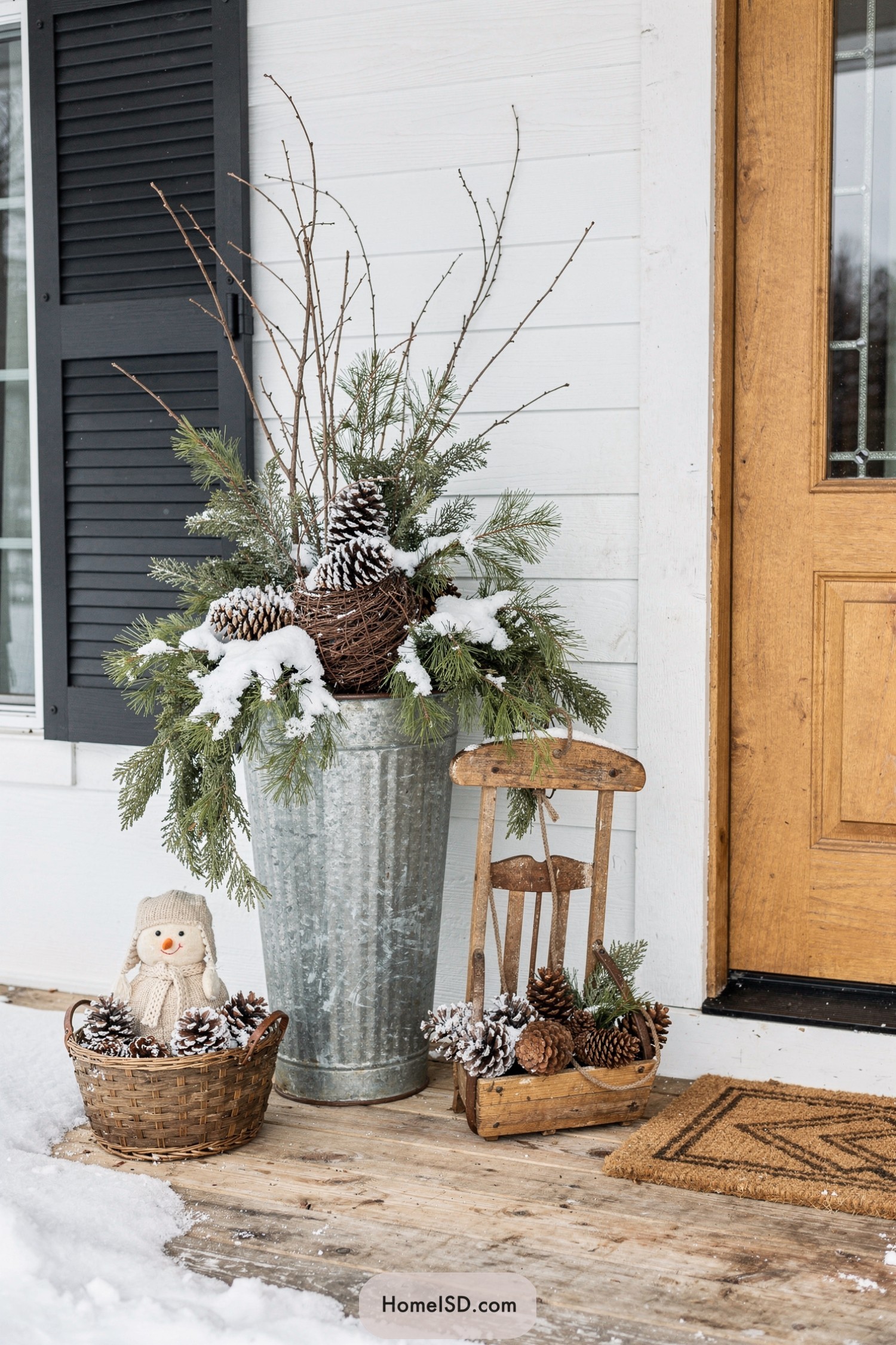 Snow-dusted evergreen and pinecone porch arrangement with rustic sled and basket decor by a wooden front door
