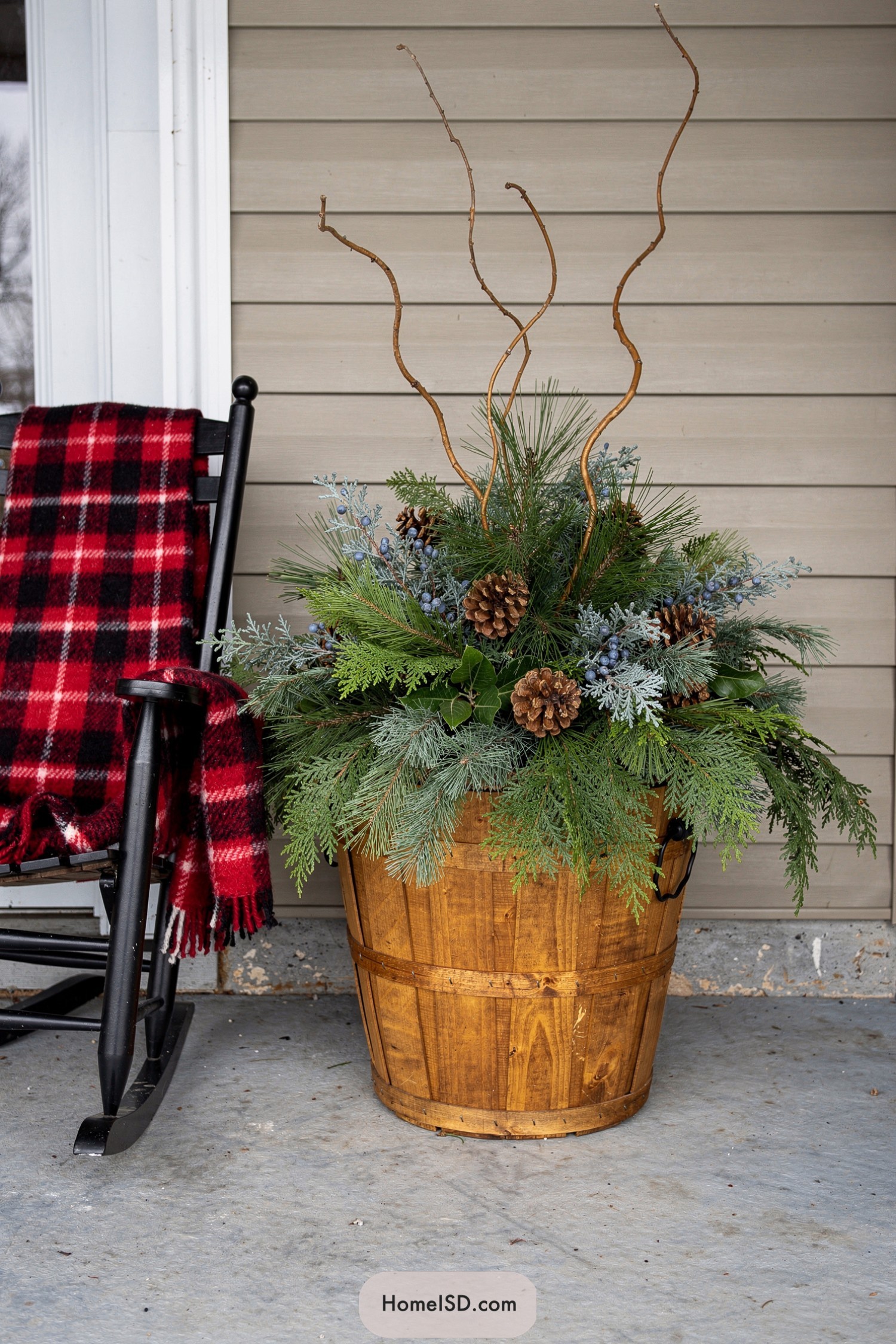Rustic wooden barrel planter with evergreens pinecones and twisting branches beside a black rocker with red plaid blanket