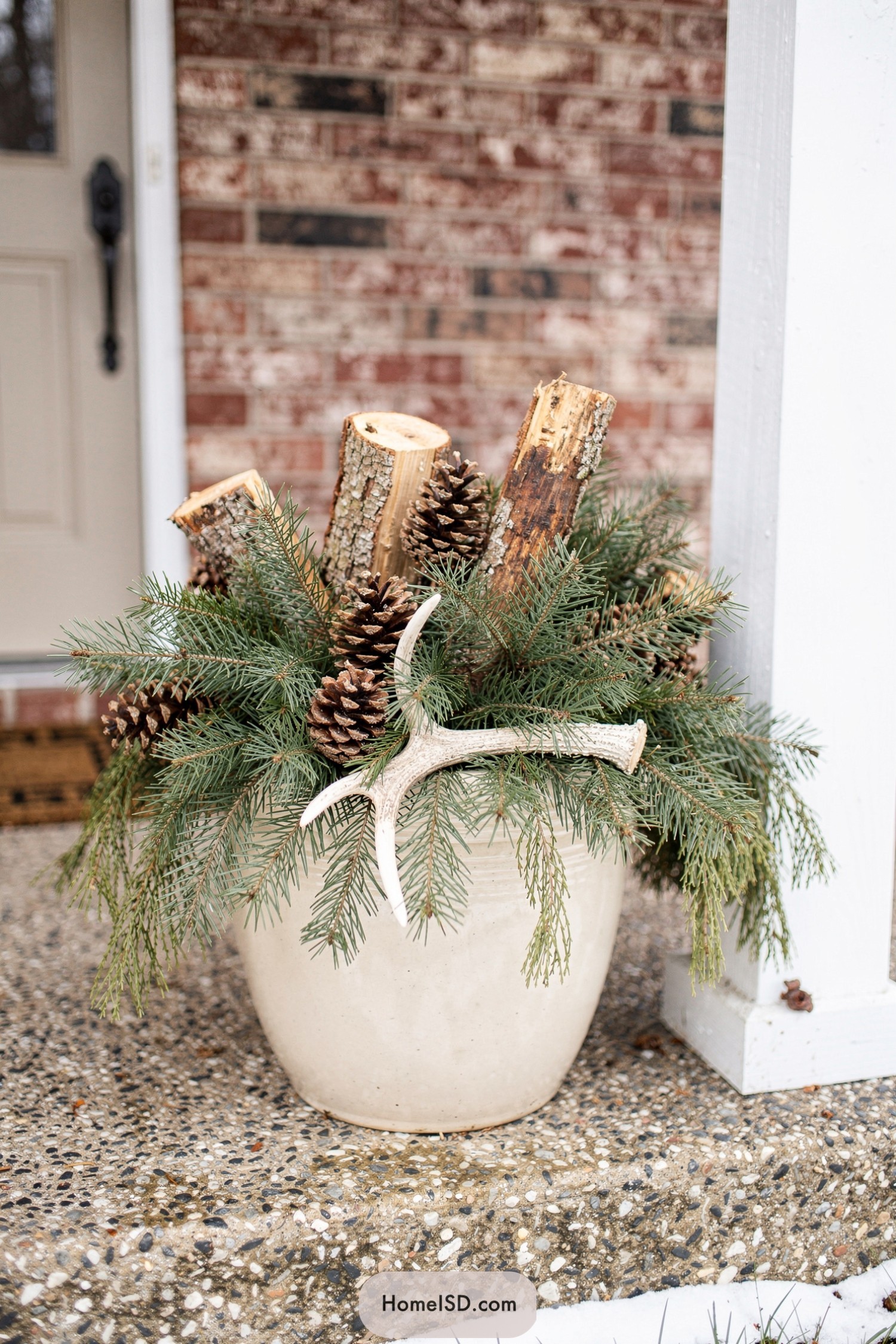 Cream planter filled with evergreens logs pinecones and antler on a front porch