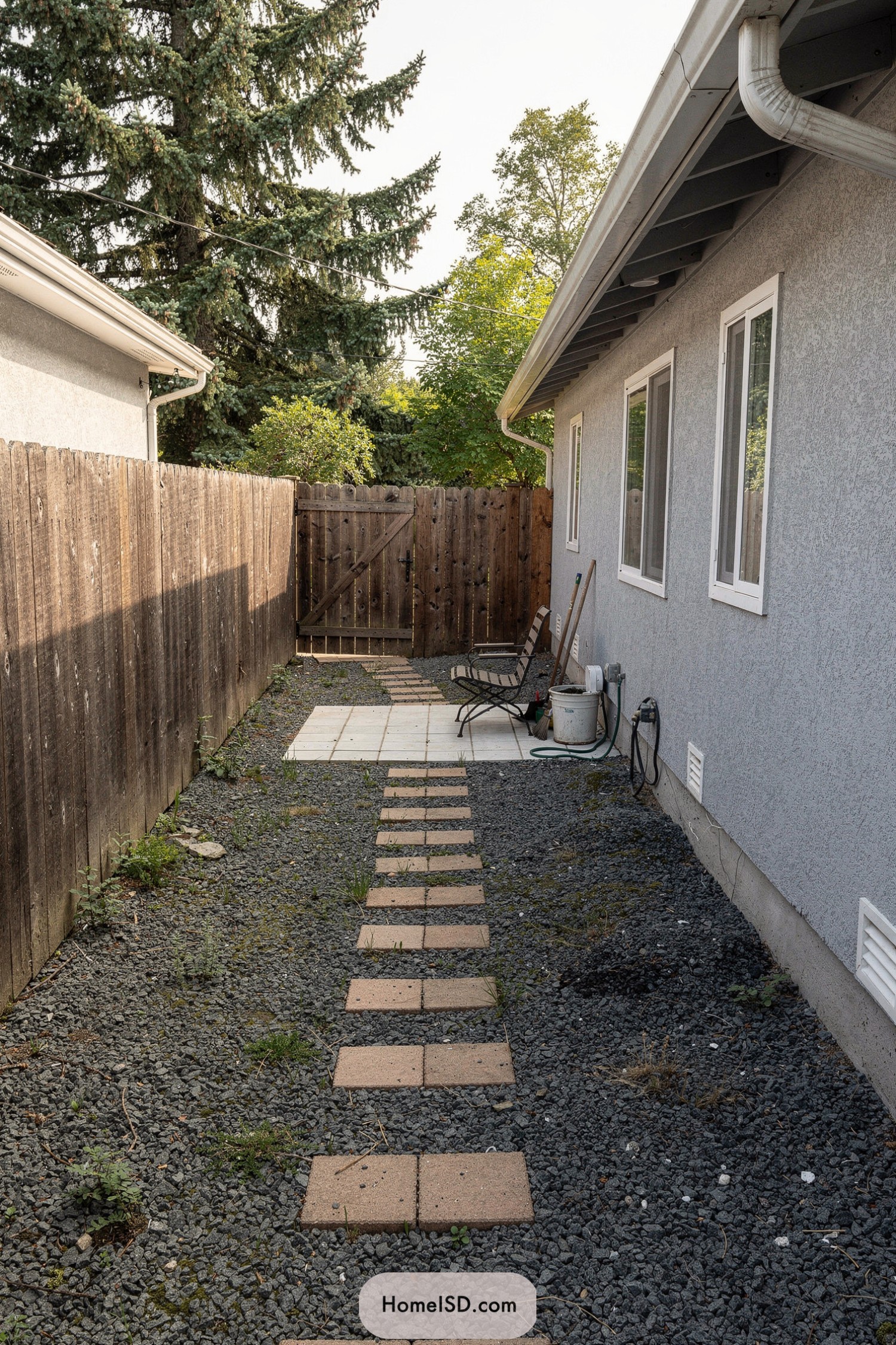 Narrow gravel side yard with stepping stones leading to a tiny tiled seating area and wooden gate