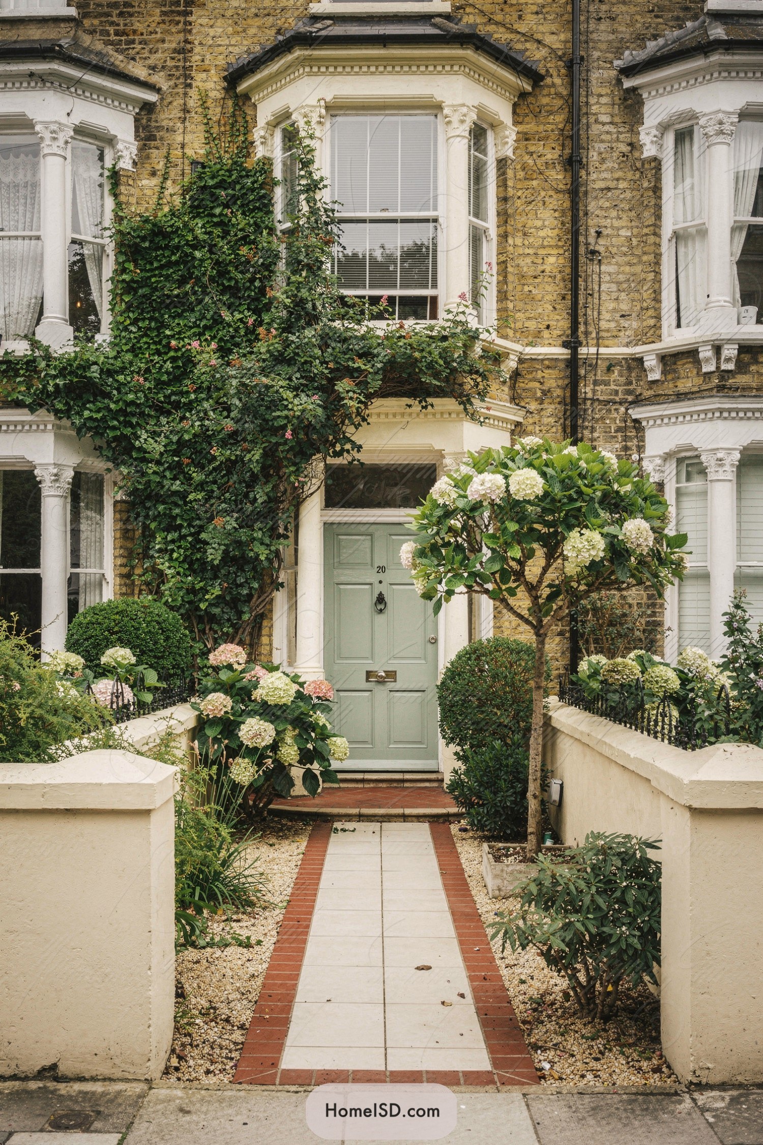 Narrow townhouse front garden with hydrangeas, clipped shrubs, and ivy framing a mint-green door along a brick-edged path