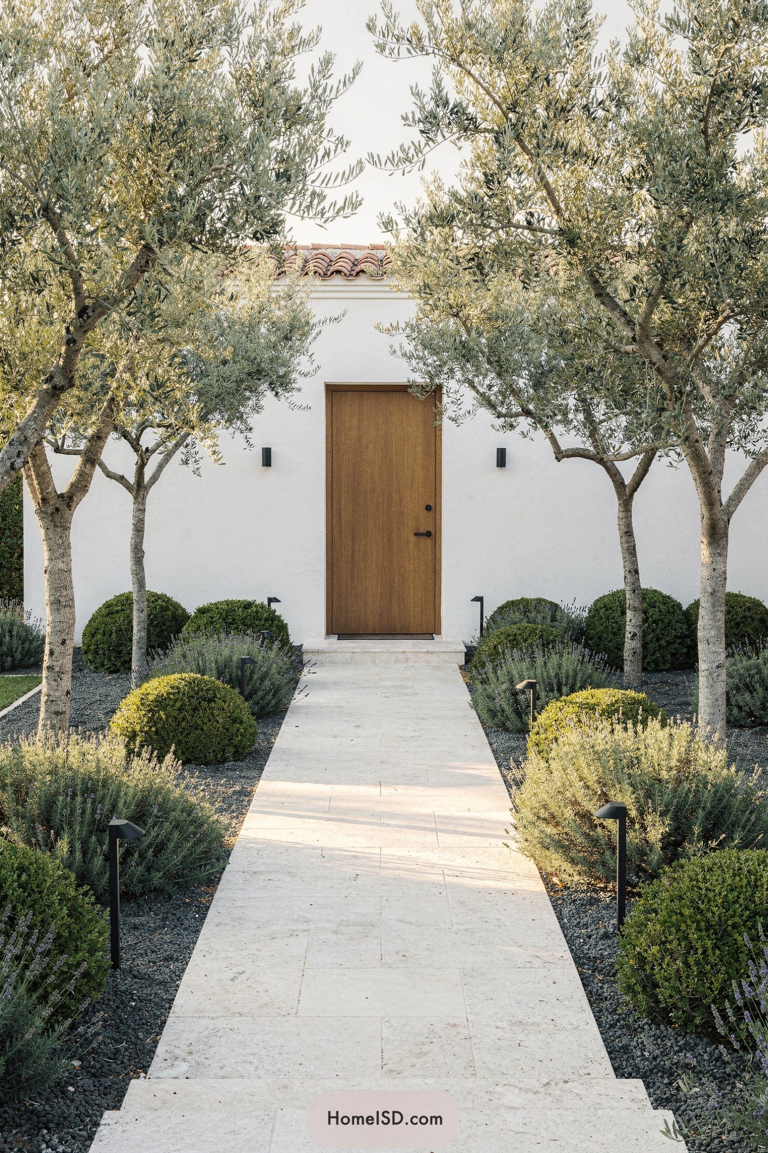 Straight limestone path flanked by olive trees and clipped shrubs leading to a simple wooden front door on a white stucco house