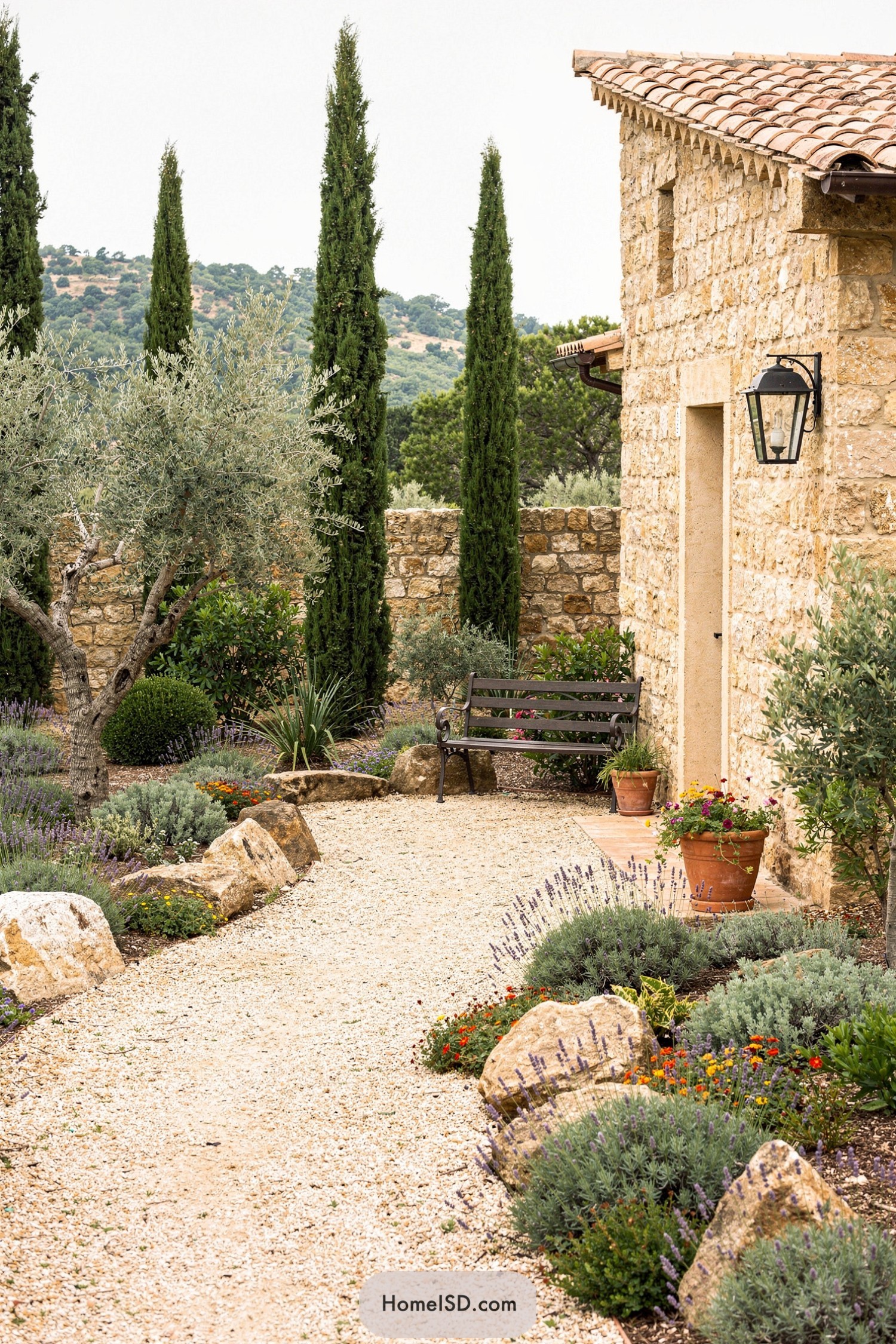 Mediterranean stone cottage with gravel path edged by boulders, lavender, and tall cypress trees leading to a bench