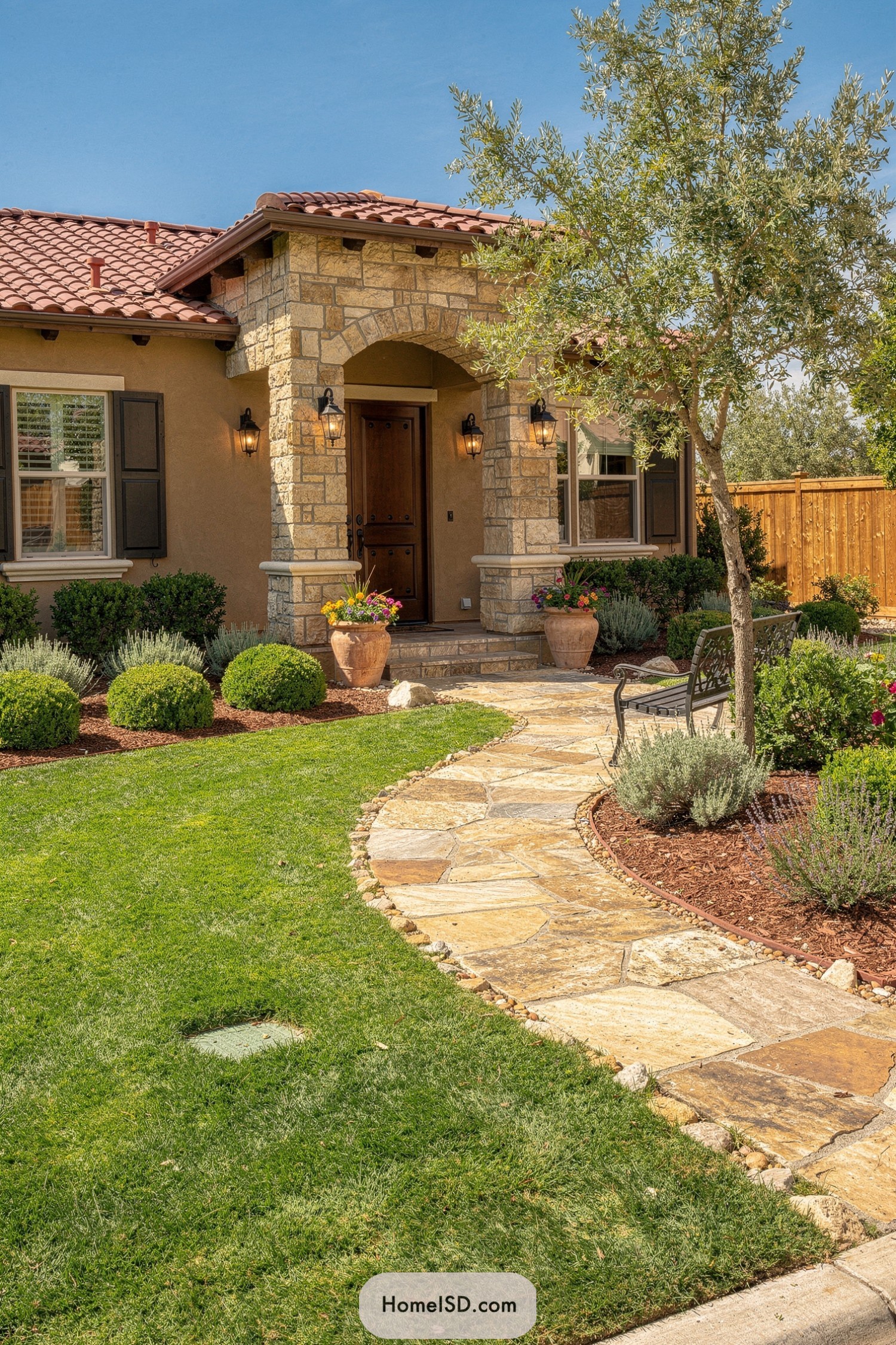 Curved flagstone path leading to a Mediterranean-style front porch with lush shrubs, potted flowers, and an olive tree