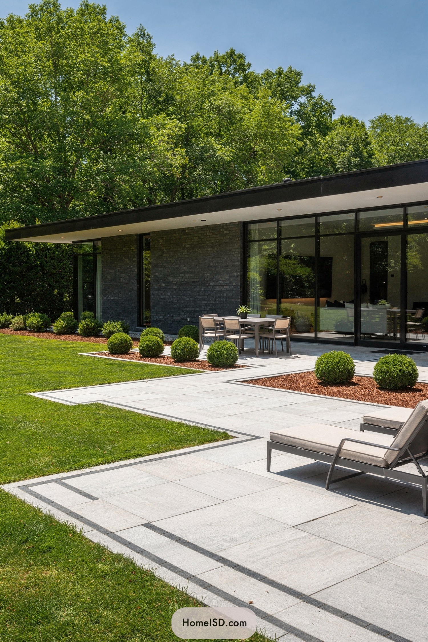 Modern hardscape patio with lawn and boxwood shrubs beside a glass-walled contemporary house