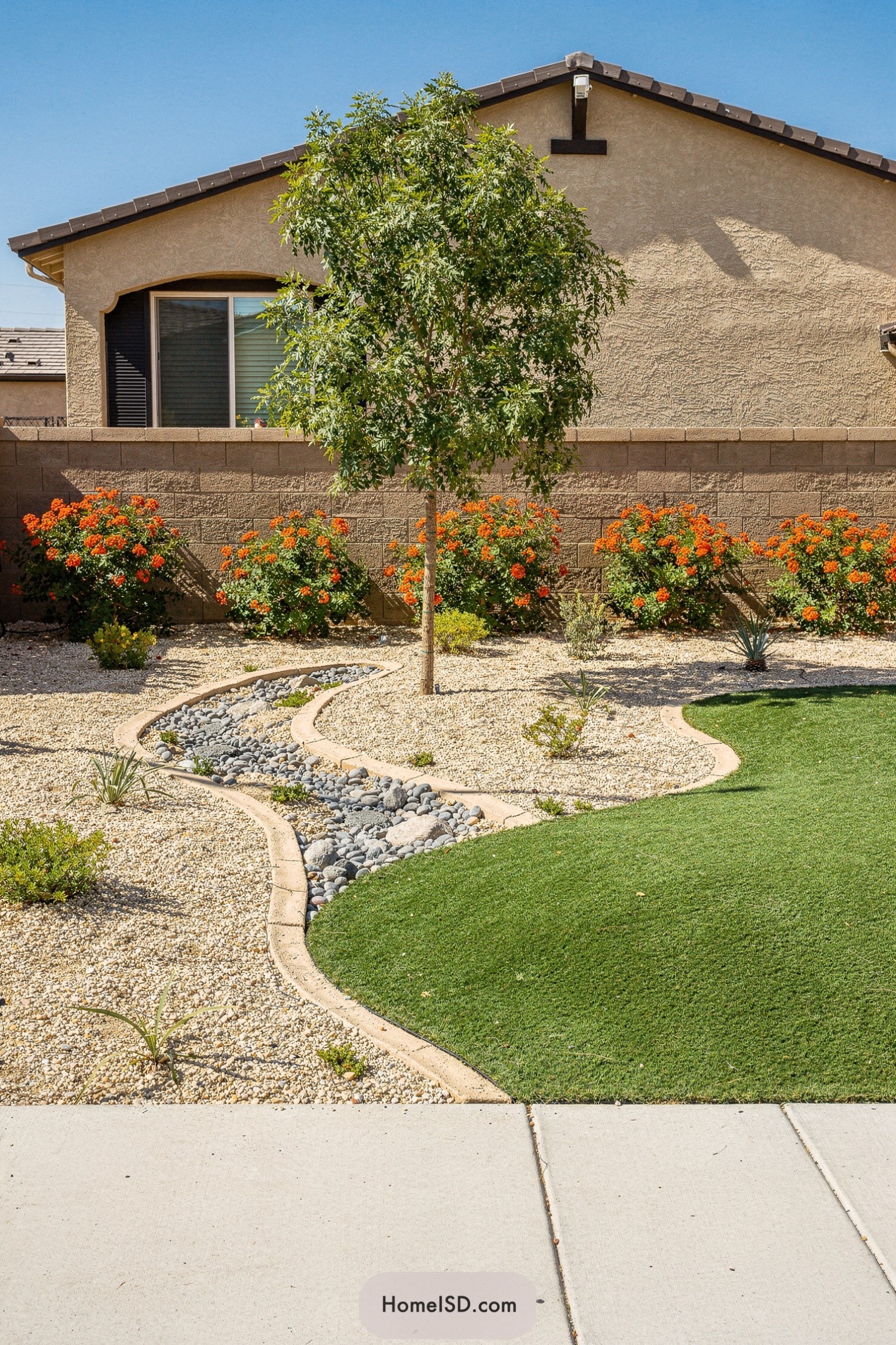 Curved dry creek bed with river rocks separating artificial turf from a gravelled desert front yard with flowering shrubs and a small tree
