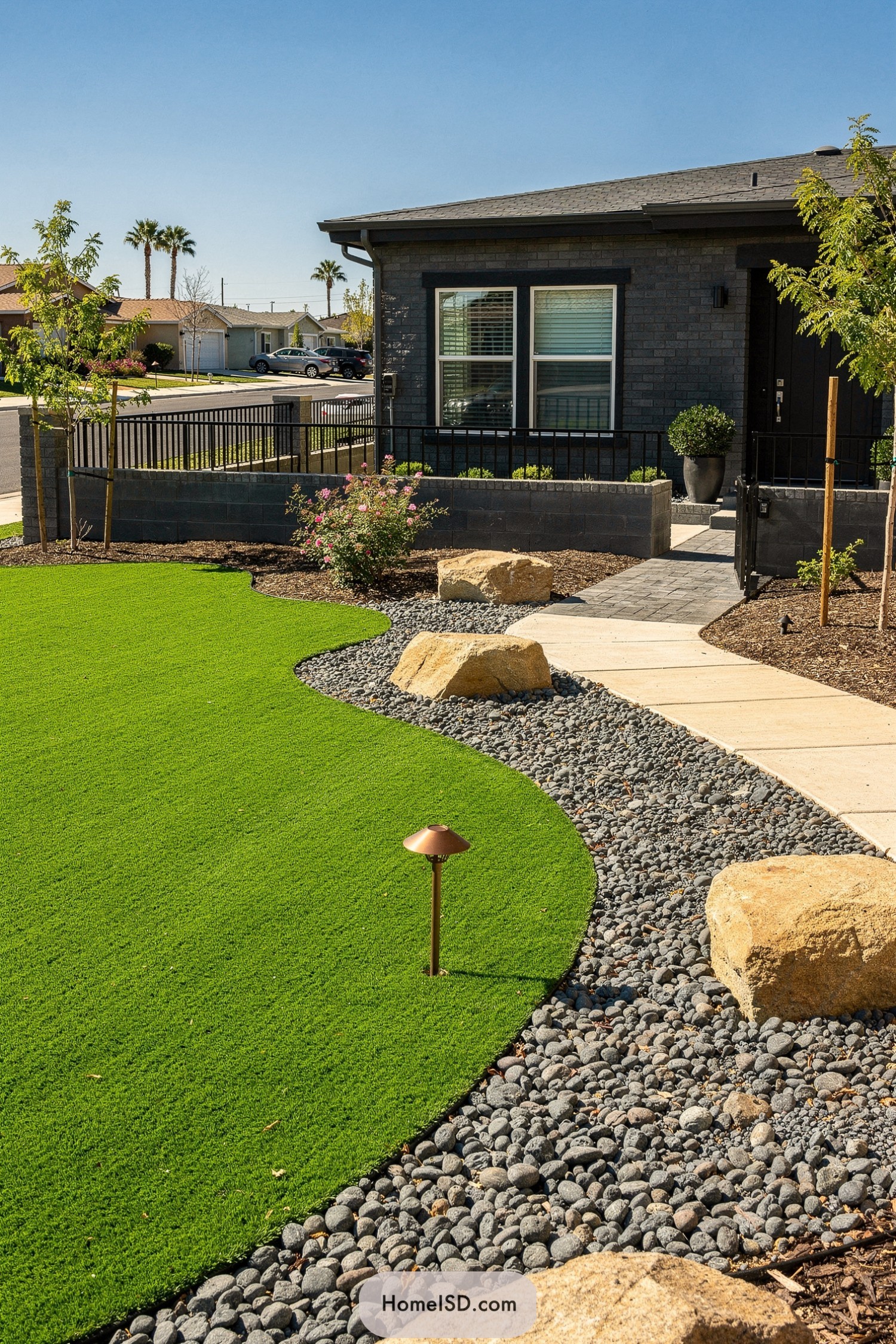 Curved artificial turf lawn with pebble border, boulders, and a paved path to a modern home