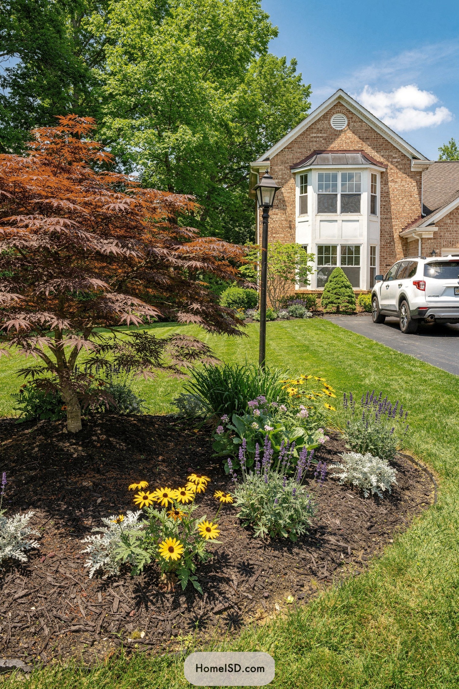 Red Japanese maple and mixed perennials in a mulched front yard bed beside a brick house and driveway