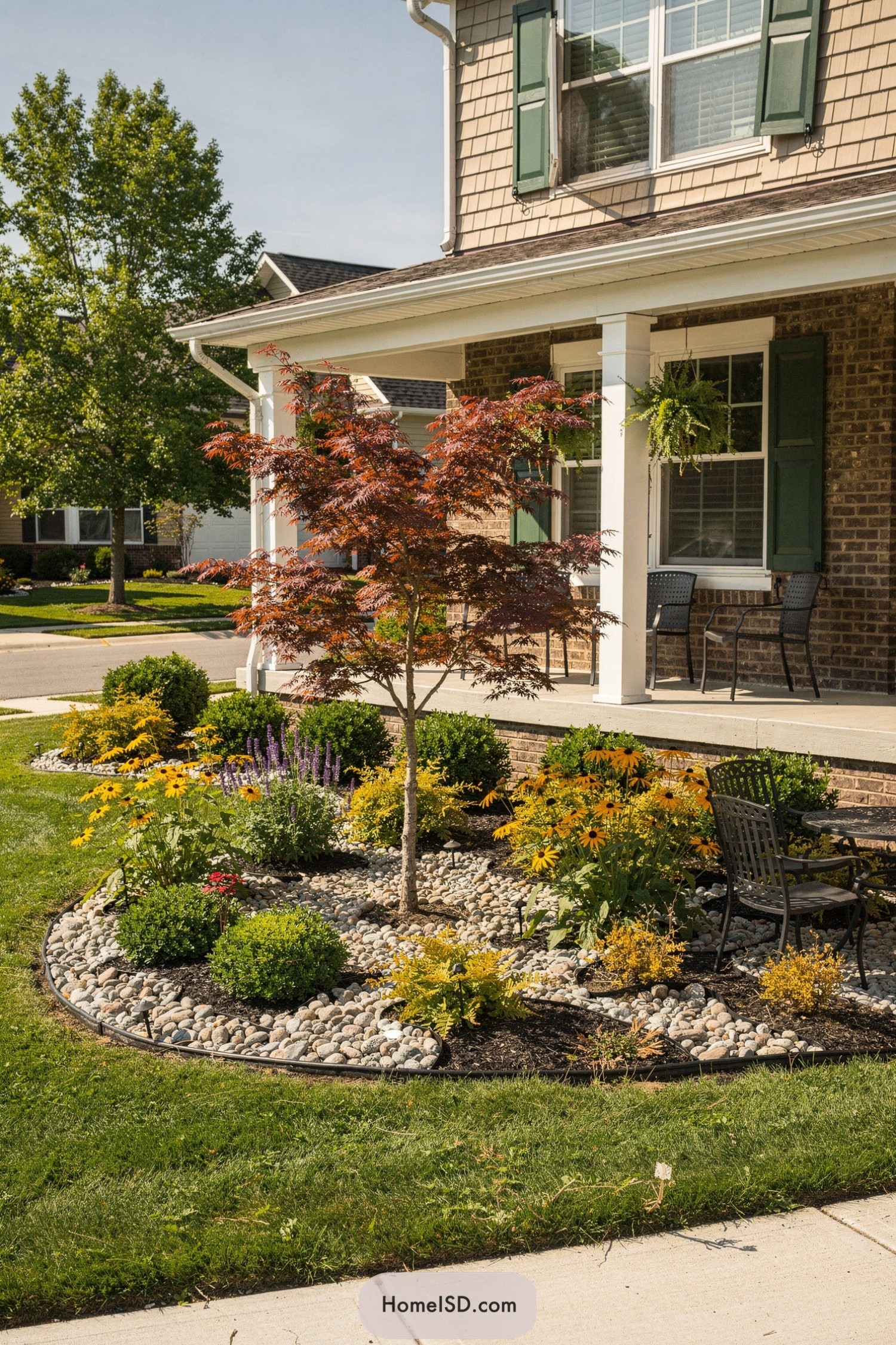 Small red maple tree in a pebble-edged front yard garden bed with colorful flowers and a small seating area on the porch
