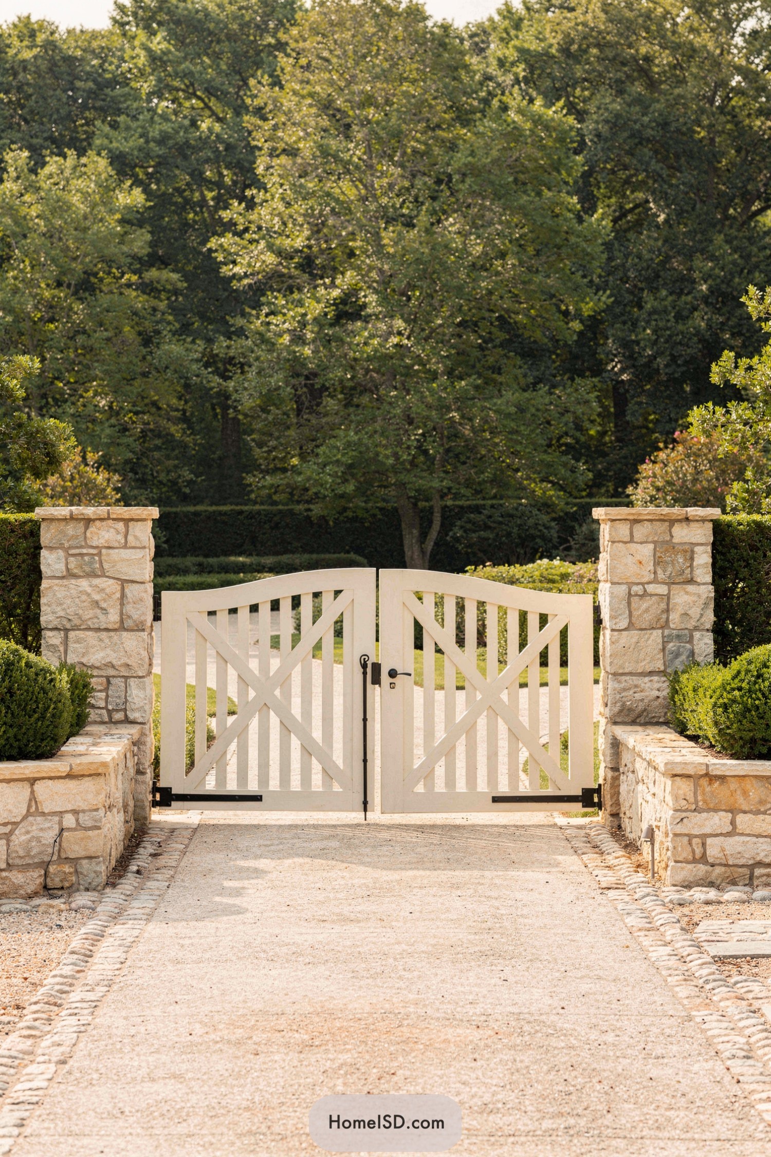 White wooden double gate with X-brace pattern between stone pillars at the end of a gravel drive