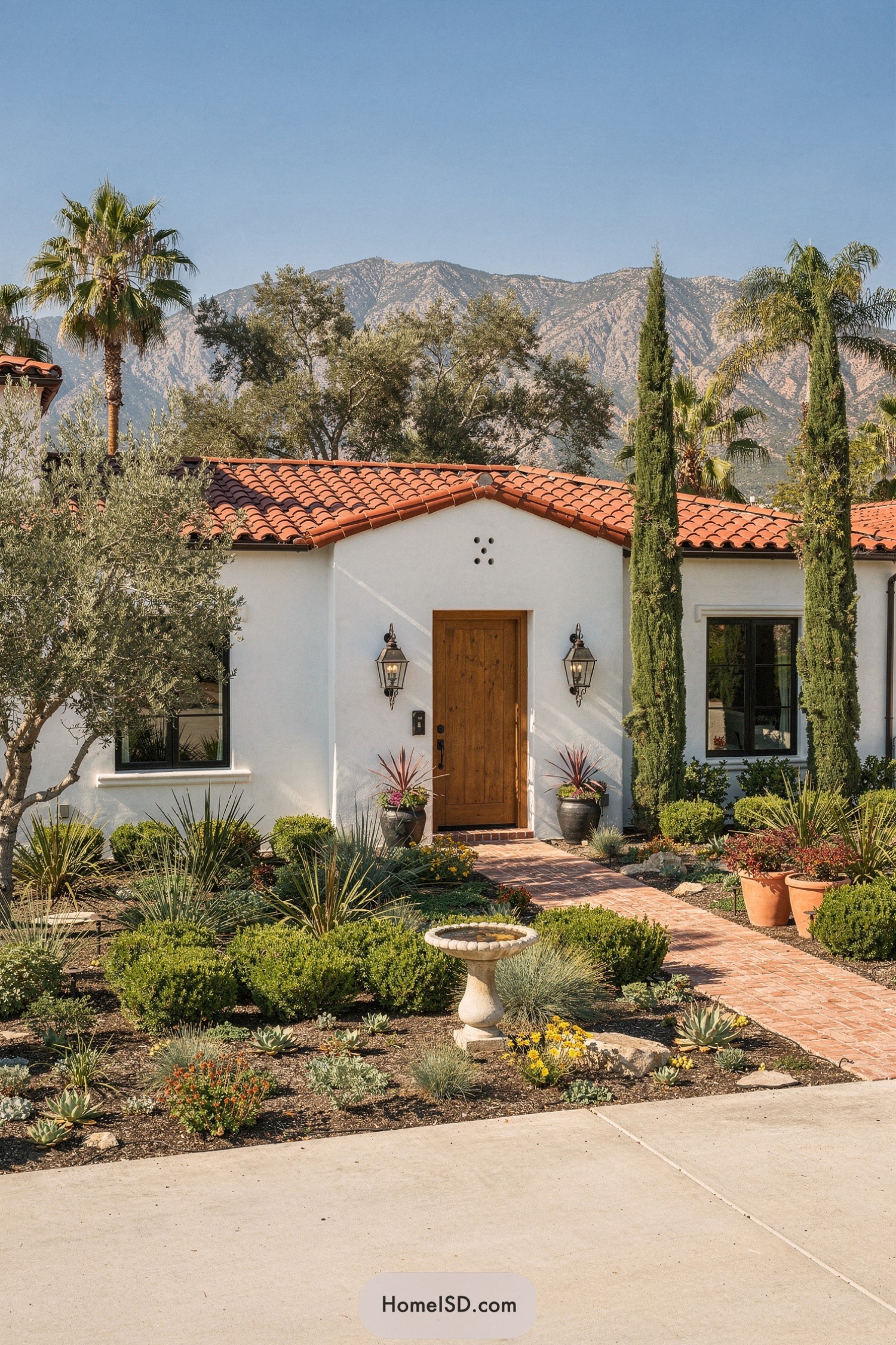 Stucco Spanish home with terracotta roof, drought-tolerant front garden, brick path, and birdbath focal point against mountain backdrop